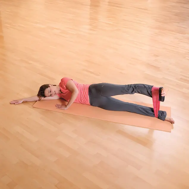 Frau liegt ausgestreckt auf der Seite auf einer Yogamatte und hat sich ein Fitnessband um die Beine gewickelt.